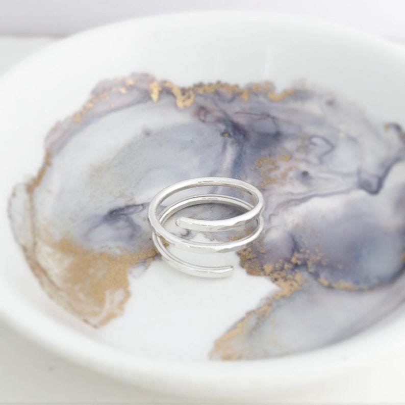 May include: A silver spiral ring with a hammered texture. The ring sits on a white and grey marbled ceramic dish with gold accents. The ring's design features multiple loops, creating a unique and modern look.
