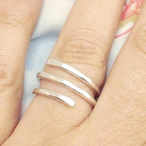 May include: A silver ring with a spiral design. The ring features three bands that wrap around the finger. The metal has a hammered texture. The ring is shown on a person's finger.