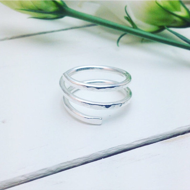 May include: A silver spiral ring with a hammered finish. The ring features an open design with three bands that coil around the finger. The ring is presented on a white surface, with a green stem and leaves visible in the background.