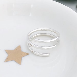 May include: A silver spiral ring with a hammered finish. The ring features three loops and is presented on a white surface, alongside a gold star-shaped cutout.