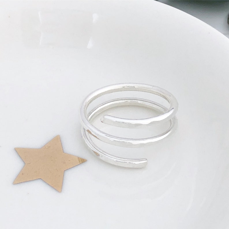 May include: A silver spiral ring with a hammered finish. The ring features three loops and is presented on a white surface, alongside a gold star-shaped cutout.