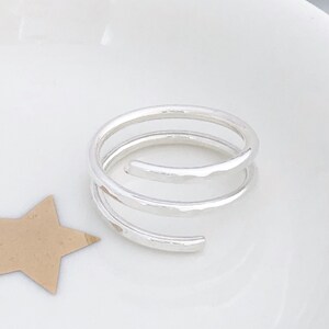 May include: A silver spiral ring with a hammered texture. The ring features a three-loop design, offering a modern aesthetic. The ring is displayed on a white surface, with a star-shaped cutout in the corner.