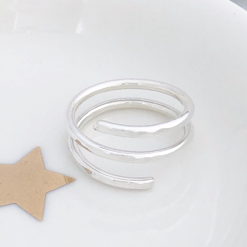 May include: A silver spiral ring with a hammered texture. The ring features a three-loop design, offering a modern aesthetic. The ring is displayed on a white surface, with a star-shaped cutout in the corner.