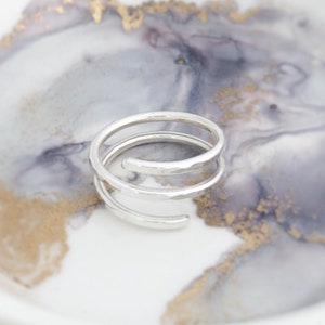 May include: A silver spiral ring with a hammered texture. The ring features three loops, forming an open design. The ring is presented on a white and grey marbled surface with gold detailing.