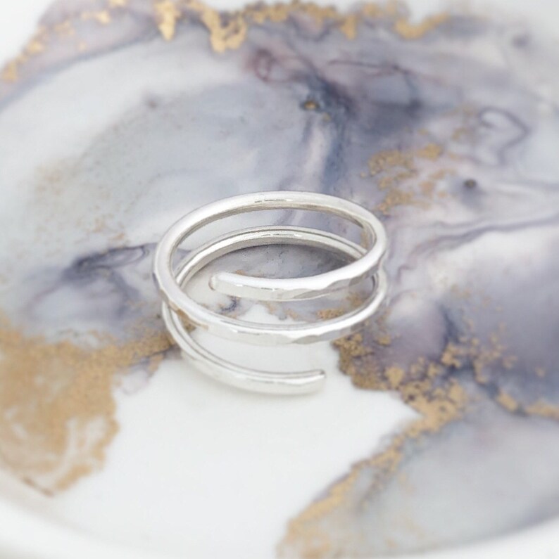 May include: A silver spiral ring with a hammered texture. The ring features three loops, forming an open design. The ring is presented on a white and grey marbled surface with gold detailing.