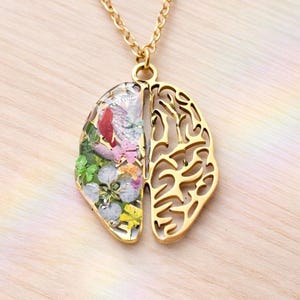 Puede incluir: Un collar con colgante dorado con un diseño en forma de cerebro. Una mitad del colgante está llena de flores secas de colores, la otra mitad es un diseño calado. El collar cuelga de una delicada cadena dorada.