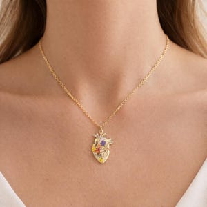 Pode incluir: Colar dourado com um pingente em forma de coração humano. O pingente em forma de coração é adornado com flores coloridas. O colar é usado ao redor do pescoço.