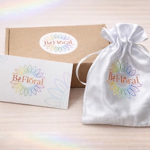 Może przedstawiać: Biały satynowy woreczek ze sznurkiem, biała karta i brązowe pudełko, wszystkie z logo "Be Floral" w tęczowych kolorach. Logo przedstawia wz&oacute;r słonecznika. Przedmioty są ułożone na jasnej powierzchni imitującej drewno.