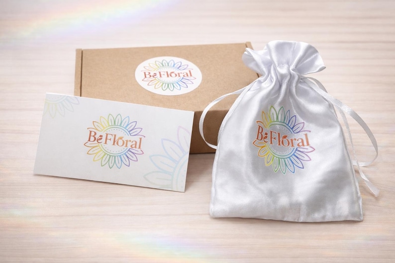 Może przedstawiać: Biały satynowy woreczek ze sznurkiem, biała karta i brązowe pudełko, wszystkie z logo "Be Floral" w tęczowych kolorach. Logo przedstawia wz&oacute;r słonecznika. Przedmioty są ułożone na jasnej powierzchni imitującej drewno.