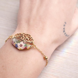 Puede incluir: Pulsera dorada con diseño floral. La pulsera tiene forma de semicírculo con un patrón de filigrana en un lado y una sección llena de flores secas de colores. La cadena es delicada con pequeños detalles de bolas.