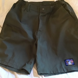 May include: Olive green shorts with an elastic waistband and button closure. Features a small patch with an eagle and flag design. The shorts have two front pockets and are made of a durable fabric.
