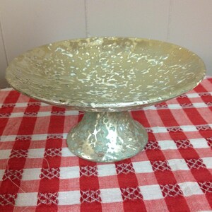 May include: A silver pedestal cake stand with a textured, speckled finish. The stand has a wide, round base and a shallow, round top.