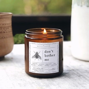 May include: A brown glass candle jar with a white label that reads "Quiet North don't bother me". The label also includes a drawing of a fly and a list of essential oils used in the candle: citronella, geranium, peppermint, pine, lavender, cinnamon, rosemary, and cedarwood.