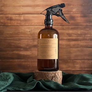 以下が含まれることがあります： 黒いスプレーノズルが付いた茶色のガラススプレーボトルで、「Multi-Purpose Household Cleaner」とラベルされています。ラベルには、ラベンダーとレモングラスのエッセンシャルオイルに関するテキストが含まれています。ボトルは木の背景に対して小さな木のスタンドにあります。