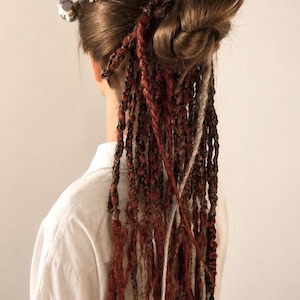 Peut inclure: Un ensemble d'extensions capillaires texturées et longues dans des tons rouge profond, brun et crème, coiffées en tresses torsadées lâches. Les cheveux sont rassemblés en chignon, ornés d'une couronne de fleurs en tissu vert pâle et blanc.