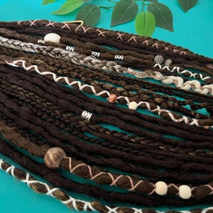Puede incluir: Una colección de rastas sintéticas de color marrón oscuro con diversas texturas, incluyendo mechones trenzados y retorcidos. El conjunto está acentuado con hilos de color rosa claro y blanco, cuentas metálicas plateadas y cuentas de madera.