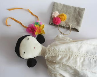 May include: A white fabric doll with black hair, wearing a white dress with lace trim. The doll is holding a yellow, pink, and red flower crown. A small brown linen bag with pom-poms is in the background.
