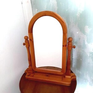 May include: A wooden vanity mirror with an arched top and a rectangular mirror. The mirror is supported by two vertical posts with decorative knobs. The base has a small shelf. The wood has a warm, natural finish.