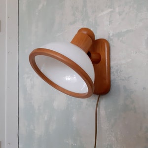Op de afbeelding: Een wandlamp in mid-century modern stijl met een witte glazen kap en een houten frame en montageplaat. De kap is omgeven door een houten ring. De lamp is bevestigd aan een muur met een lichtblauwe en witte verf.