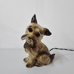 Puede incluir: Lámpara de cerámica con forma de perro, en tonos marrón y beige. La lámpara presenta un rostro detallado con ojos grandes y un hocico largo. El perro está sentado, con un cable eléctrico marrón que se extiende desde la parte trasera. Un artículo decorativo.
