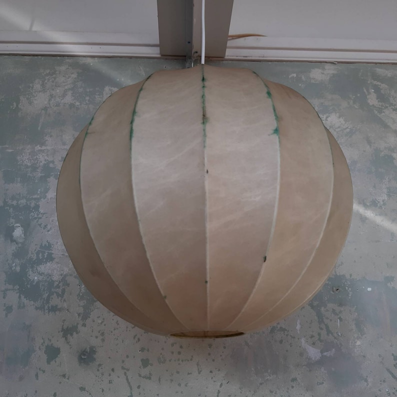 May include: A white, round, pleated lampshade hangs from the ceiling. The lampshade has a slightly textured surface and is made of a thin, flexible material.