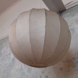 May include: A white, round, pleated lampshade hangs from the ceiling. The lampshade is made of a soft, fabric-like material and has a slightly textured surface.