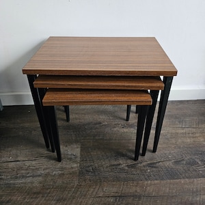 May include: A set of three nesting tables with a dark wood grain finish on the square tabletops. The tables have black metal legs. The largest table is at the top, with the smaller tables nested underneath.