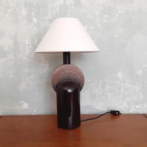 May include: A contemporary table lamp featuring a white lampshade, a dark brown spherical centre, and a glossy, dark brown base. The lamp is plugged into a wall outlet. The lamp is on a wooden surface.