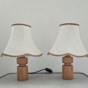 Pu&ograve; includere: Due lampade da tavolo in legno con paralumi in lino bianco e bordatura marrone. Le lampade hanno una finitura in legno naturale e un design semplice e cilindrico.