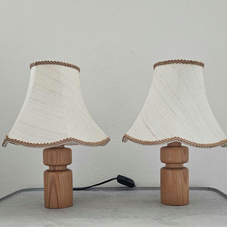 Pu&ograve; includere: Due lampade da tavolo in legno con paralumi in lino bianco e bordatura marrone. Le lampade hanno una finitura in legno naturale e un design semplice e cilindrico.