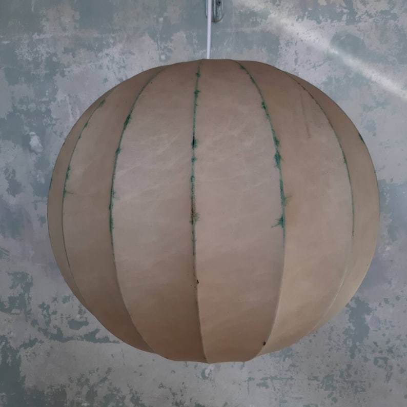 May include: A beige, round, fabric lampshade with vertical seams. The lampshade is suspended from a white cord.