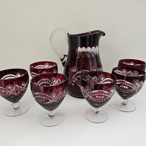 May include: A ruby red crystal glassware set, comprising a jug and six stemmed glasses. The glassware has detailed cut patterns and clear glass stems and handle. The set is displayed on a white surface.