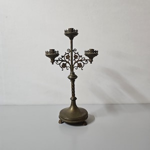 Könnte beinhalten: Ein antiker Messing-Kandelaber mit drei Kerzenhaltern. Der zentrale Stiel hat ein gedrehtes Design, und die Arme weisen florale Verzierungen auf. Der Sockel ist rund mit kleinen Klauenfüßen. Der Kandelaber ist ein Vintage-Dekorationsartikel.