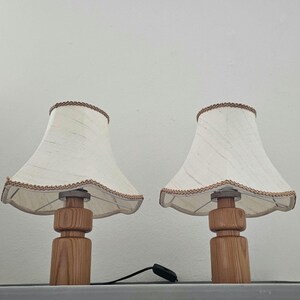 Pu&ograve; includere: Due lampade da tavolo in legno con paralumi in tessuto bianco e bordatura marrone. I paralumi hanno un bordo a festoni.