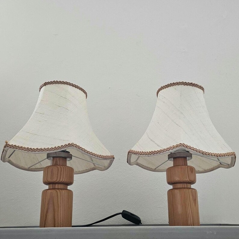 Pu&ograve; includere: Due lampade da tavolo in legno con paralumi in tessuto bianco e bordatura marrone. I paralumi hanno un bordo a festoni.