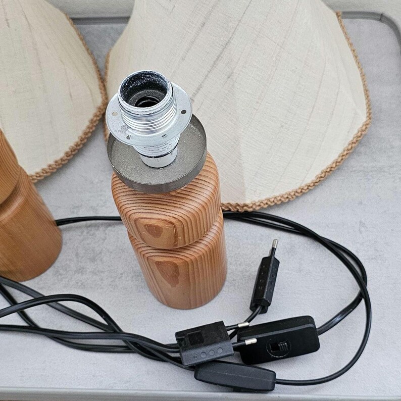 Pu&ograve; includere: Due lampade da tavolo in legno con paralumi in tessuto bianco. Le lampade hanno un design semplice con una base rotonda e un corpo cilindrico. Le lampade sono collegate a un cavo elettrico nero con un interruttore.