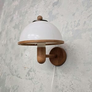 Peut inclure: Une applique murale avec un abat-jour blanc en forme de dôme et une base en bois. La lampe présente une garniture en bois autour de l'abat-jour et un petit bouton en bois sur le dessus. Le luminaire est fixé au mur avec une plaque en bois ronde.