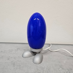 May include: A modern table lamp with a vibrant blue, egg-shaped shade. The lamp sits on a silver base with two foot-shaped supports. A white power cord extends from the base.