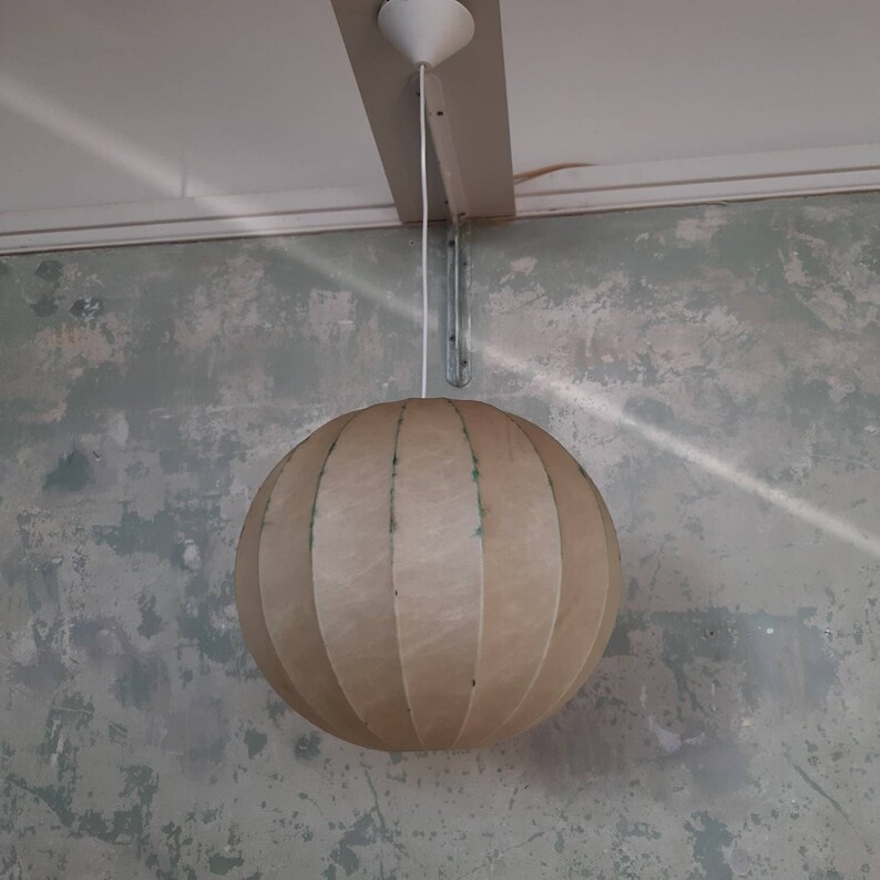 May include: A beige, round, hanging lampshade with vertical lines. The lampshade is made of a textured material and is hanging from a white cord.