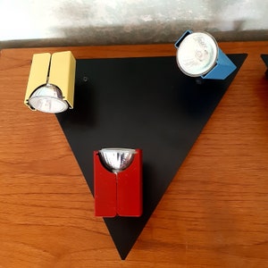 May include: A contemporary lighting fixture with three spotlights in yellow, blue, and red. The lights are mounted on a black triangular base, set against a wooden surface. Each spotlight has a silver bulb.