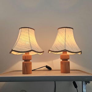 Pu&ograve; includere: Due lampade da tavolo in legno con paralumi in tessuto bianco. Le lampade hanno un design semplice e cilindrico e sono accese, illuminando la stanza.