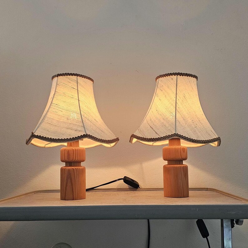 Pu&ograve; includere: Due lampade da tavolo in legno con paralumi in tessuto bianco. Le lampade hanno un design semplice e cilindrico e sono accese, illuminando la stanza.