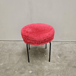 Puede incluir: Un taburete redondo con un cojín de asiento de piel sintética de color rosa intenso. El asiento está sostenido por cuatro patas de metal negro, finas y rectas, que se extienden hacia abajo desde la base del cojín, proporcionando estabilidad.