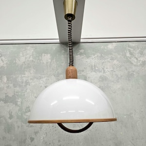 May include: A white pendant light with a wooden base and a coiled cord. The light has a white, dome-shaped shade with a wooden trim.
