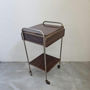 May include: A brown wooden two-tiered rolling cart with a metal frame. The top tier has a drawer and the bottom tier is a shelf.