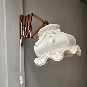 Könnte beinhalten: Eine Wandleuchte mit einem hölzernen Ziehharmonika-Arm und einem weißen Stoffschirm. Der Schirm hat einen gerüschten Rand und einen Kordelzugverschluss. Die Lampe ist an einer grauen Wand befestigt und hat ein weißes Kabel mit einem Ein-/Ausschalter.