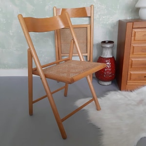 May include: A light-colored wooden folding chair with a woven seat and back. The chair is in a room with a light green wall, a red and white vase, and a wooden cabinet. Another folding chair is behind it.