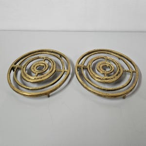 Può includere: Due sottopentola color ottone con un design a spirale. Ogni sottopentola presenta una serie di cerchi concentrici, creando un motivo strutturato. I sottopentola ovali hanno piccoli piedini per la stabilità.