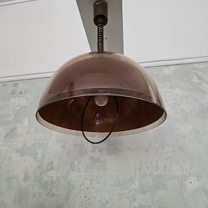 May include: A copper-colored, dome-shaped pendant light fixture. The light has a clear, rounded shade with a copper-colored interior and a black wire ring around the bulb. The fixture is suspended from the ceiling by a metal rod and a spring.