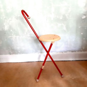 May include: A vintage red metal folding chair with a beige seat. The chair has a curved back and a round seat with several small holes. The legs form an X shape, and the chair has small white feet.